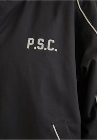 Jachetă din țesătură închisă, cu o margine alb contrastantă și logo-ul brodat "P.S.C.". Textură netedă, cu un croi structurat și cusături detaliate.
