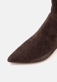 Gros plan d'une botte en daim brun foncé à bout pointu sur un fond neutre.
