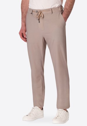 Uomo che indossa pantaloni beige con coulisse e sneakers bianche con le mani nelle tasche su uno sfondo semplice.