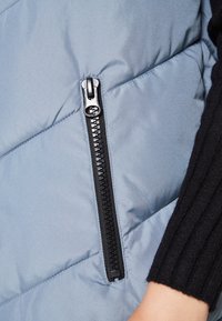 Nahaufnahme eines schwarzen Reißverschlusses an einer hellblauen gesteppte Jacke mit dem Arm einer Person in einem schwarzen gerippten Ärmel in der Nähe.