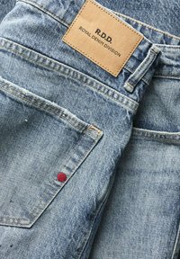 Blaue Denim-Jeans mit einem Lederpatch mit der Aufschrift "R.D.D. Royal Denim Division" und sichtbarer Tasche mit rotem Nieten-Detail.