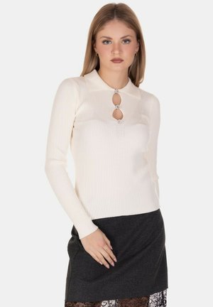 Una donna con capelli biondi lisci indossa un top a maniche lunghe color crema a coste con tre bottoni gioiello e una gonna scura con bordo di pizzo.