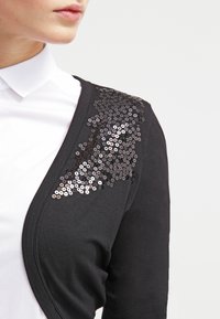 Donna che indossa un cardigan nero con paillettes nere sulle spalle su una camicia bianca con colletto, mostrata dalla gola fino a metà torso.
