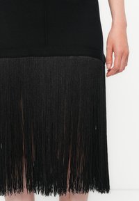 Svart kjole med en tettsittende overdel og frynsete svart kant. Frynsene har en tekstur som skaper bevegelse. Materialet er mykt og elastisk.