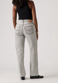 Femme portant un jean gris clair droit et un haut noir sans manches, debout dos à la caméra sur un fond uni.