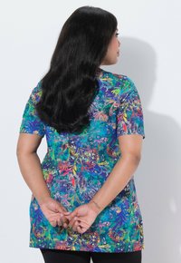 Kvinde med langt sort hår står med hænderne sammenlagt bag ryggen iført en farverig blomsterprintet kortærmet top mod en ensfarvet hvid baggrund.