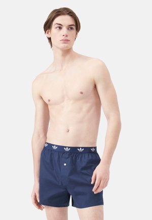 Junger Mann mit heller Haut und braunen Haaren, trägt dunkelblaue Adidas-Boxershorts, steht vor einem schlichten hellen Hintergrund.