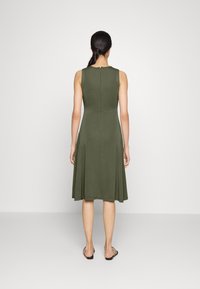 Kobieta stoi tyłem, ubrana w bezrękawnikową oliwkową sukienkę, która rozszerza się na dole, a jej włosy są ułożone w niski kucyk.