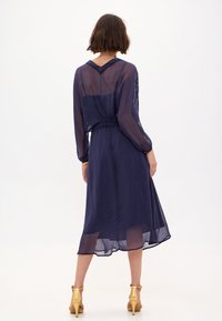 Robe bleu marine à manches longues en tissu transparent, avec une taille cintrée et un ourlet au genou. Sandales à talons beiges assorties et accents dorés visibles.