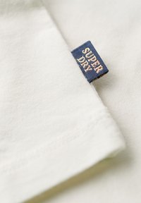 Witte katoenen t-shirt met een opgevouwen zoom en een blauw label met de tekst "SUPERDRY" in gouden letters, die een gladde structuur toont.
