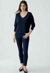Femme portant un pull V-nec marine, un pantalon skinny assorti, des ballerines pointues noires et des boucles d'oreilles créoles, se tenant devant un fond clair.