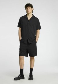 Camicia nera a maniche corte con bottoni e pantaloni corti abbinati. Tessuto liscio, vestibilità rilassata e design minimalista. Mocassini neri e calzini completano il look.