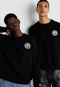 Sorte sweatshirt laget av myk stoff med rund hals. Forsiden viser en hvit brodert logo. Modellen til venstre har en cropped passform.