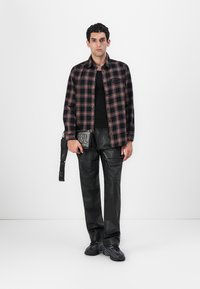 Camicia a quadri nera, rossa e bianca, abbinata a pantaloni in pelle neri. Il modello tiene una borsa nera con logo e tracolla bianchi.