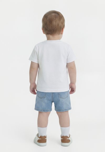 Νήπιο που φοράει λευκό μπλουζάκι, denim σορτς, λευκές κάλτσες και καφέ-λευκά αθλητικά παπούτσια, στέκεται γυρισμένο με την πλάτη σε λευκό φόντο.