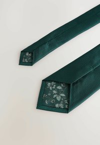 Grøn slips med et struktureret, diamantmønster. Det har en blomsterforet indvendig sektion, som viser en kontrasterende design detalje ved spidsen.