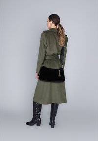 Donna con capelli lunghi raccolti, che indossa un lungo cappotto verde oliva e stivali neri fino al ginocchio, con una borsa nera dietro la schiena.