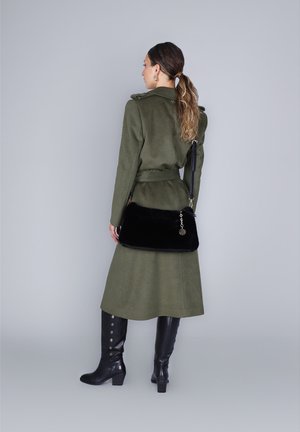 Donna con capelli lunghi raccolti, che indossa un lungo cappotto verde oliva e stivali neri fino al ginocchio, con una borsa nera dietro la schiena.