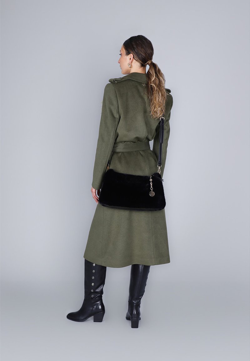 Donna con capelli lunghi raccolti, che indossa un lungo cappotto verde oliva e stivali neri fino al ginocchio, con una borsa nera dietro la schiena.