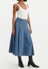 Jupe en denim en A avec une taille haute, dotée d'une fermeture à boutons sur le devant et de surpiqûres contrastantes. Portée avec un débardeur blanc et des bottes noires.