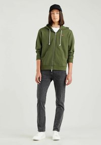 Felpa con zip verde oliva e lacci bianchi, tasche frontali, abbinata a jeans scuri e sneakers bianche. Un cappellino nero completa l'outfit.