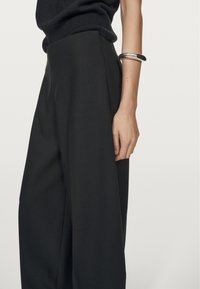 Sorte wide-leg bukser lavet af glat stof, med høj talje og subtile plisseringer. Et sølvarmbånd bæres om håndleddet.