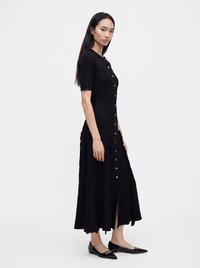 Γυναίκα με μακριά σκούρα μαλλιά φοράει ένα μαύρο midi φόρεμα με πιέτες και κουμπιά μπροστά και μαύρες μυτερές μπαλαρίνες, στέκεται πλάγια σε λευκό φόντο.