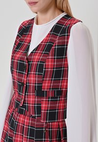 Gilet a quadri rosso e nero con design aderente, caratterizzato da bottoni e due tasche, indossato sopra una blusa bianca a maniche lunghe.