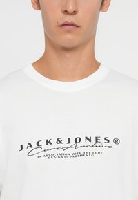 Λευκό βαμβακερό T-shirt με μαύρο γραφικό τύπωμα που περιλαμβάνει το κείμενο "JACK & JONES" και "Σε συνεργασία με το Τμήμα Σχεδιασμού Core."