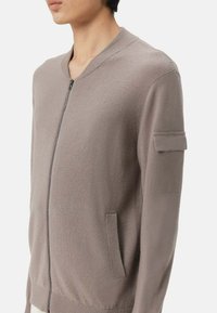 Ljusbrun zip-up cardigan med en ribbad krage, sidofickor och en texturerad yta. Har en ficka på vänster ärm.
