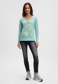 Hellblaugrünes Langarm-T-Shirt mit grafischem Text in verschiedenen Farben. Kombiniert mit dunklen Jeans und grauen Sneakers mit mintfarbenen Akzenten.