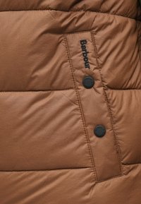 Barna párnázott kabát sima textúrával, fekete gombos rögzítéssel és a "Barbour" hímzett logójával az oldalán.