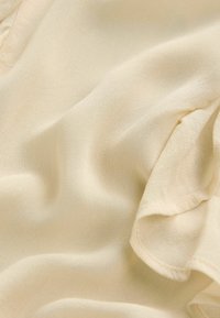 Tissu doux de couleur crème avec des plis visibles et une texture subtile, montrant un détail de bord froncé sur le côté droit.
