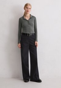 Kobieta z blond włosami w niskim kucyku, ubrana w szary, prążkowany, długi rękaw polo, czarne szerokie spodnie, czarny pasek i czarne buty, stoi przed białą ścianą.