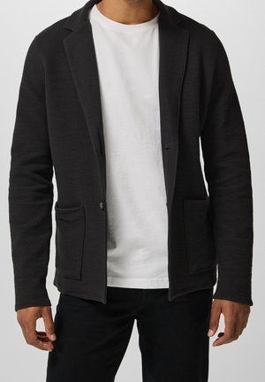 Mann trägt einen schwarzen strukturierten Blazer über einem weißen T-Shirt und schwarzen Hosen, steht mit entspannten Armen an den Seiten.