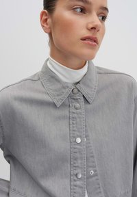Gray denim shirt with a collar, snap buttons, and visible stitching. Worn over a white turtleneck, featuring a soft texture.