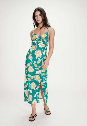 Femme debout portant une robe mi-longue bleu sarcelle à imprimé floral jaune, bretelles spaghetti, fente avant, et sandales noires à lanières.