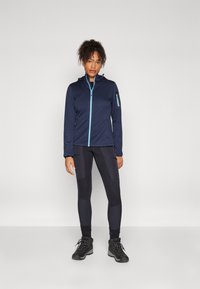 Giacca softshell blu navy con zip azzurro chiaro, abbinata a leggings neri con toppe testurizzate e robuste scarpe da escursionismo nere.