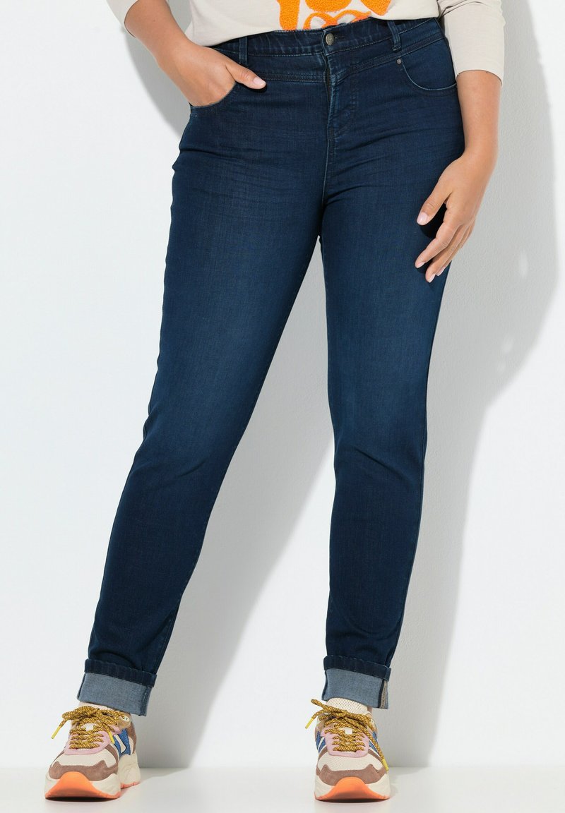 Jean skinny en denim foncé avec taille haute et ourlet retroussé, associés à des baskets multicolores avec des accents beiges, roses et bleus.