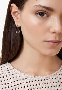 Orecchini a cerchio in argento con una texture torcillata, posizionati su un top leggero intrecciato. Visione ravvicinata che mette in evidenza i dettagli dell'orecchino e del viso.