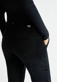 Pantaloni in corduroy neri con una texture liscia e un design aderente, caratterizzati da un logo discreto vicino alla vita e tasche frontali.