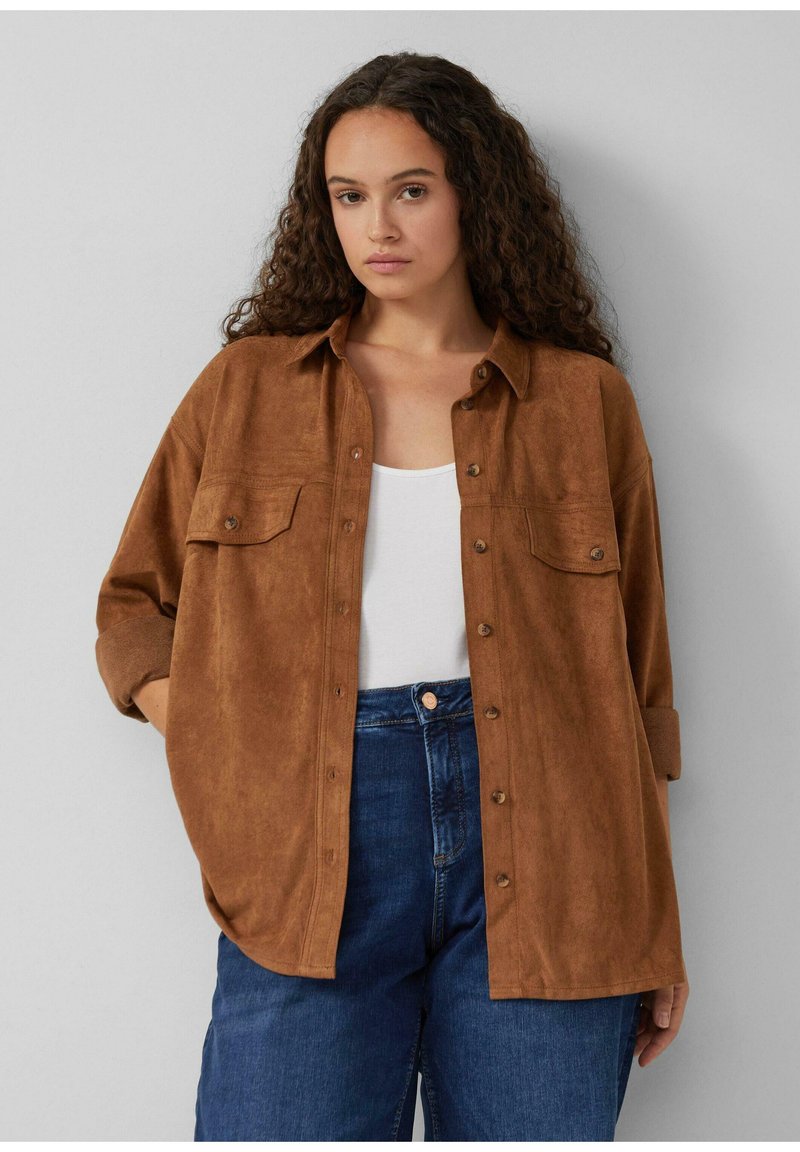 Frau mit langen lockigen Haaren, die ein braunes Button-up-Shirt über einem weißen Oberteil und blauen Jeans trägt, steht vor einer schlichten Wand.