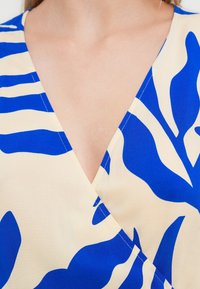 Femme portant un haut à col en V crème et bleu à motif, montrant le haut de la poitrine et l'encolure.