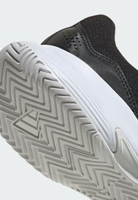 Czarne buty sportowe z fakturowaną cholewką, białą gumową podeszwą z zygzakowatym wzorem oraz małym logo na podeszwie.