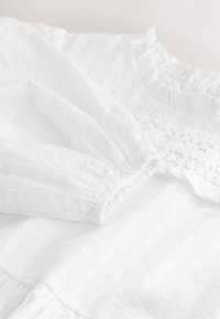 White dress featuring textured fabric with a subtle diamond pattern, elasticized sleeves, and delicate crochet detailing along the bodice.