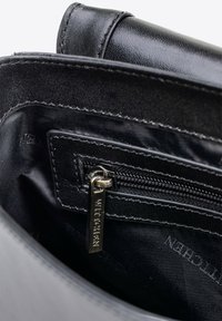 Interior de un bolso sintético negro que presenta un bolsillo con cremallera y una etiqueta de metal plateado, textura suave y detalles de costura.