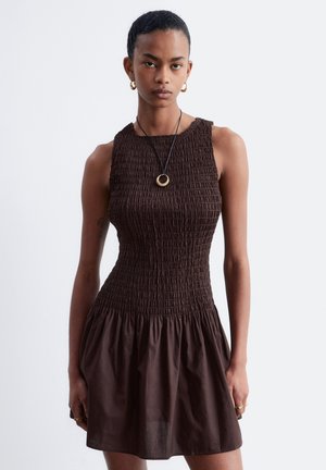 Personne mince portant une robe smockée sans manches marron foncé, des boucles d'oreilles créoles dorées et un collier avec pendentif rond, debout devant un fond uni.
