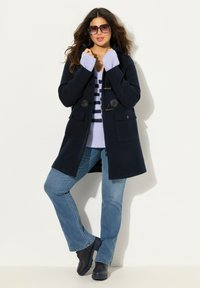 Cappotto in lana blu con bottoni a toggles e tasche frontali, indossato sopra un maglione a righe viola chiaro. Jeans blu e scarpe nere completano l'outfit.