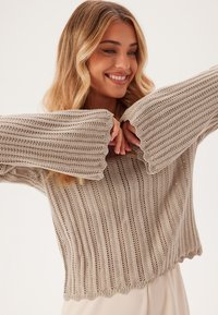 Maglione lavorato a maglia di color marrone chiaro, con maniche larghe a coste e orlo a scallop. Presenta un design testurizzato a maglia aperta con righe verticali.