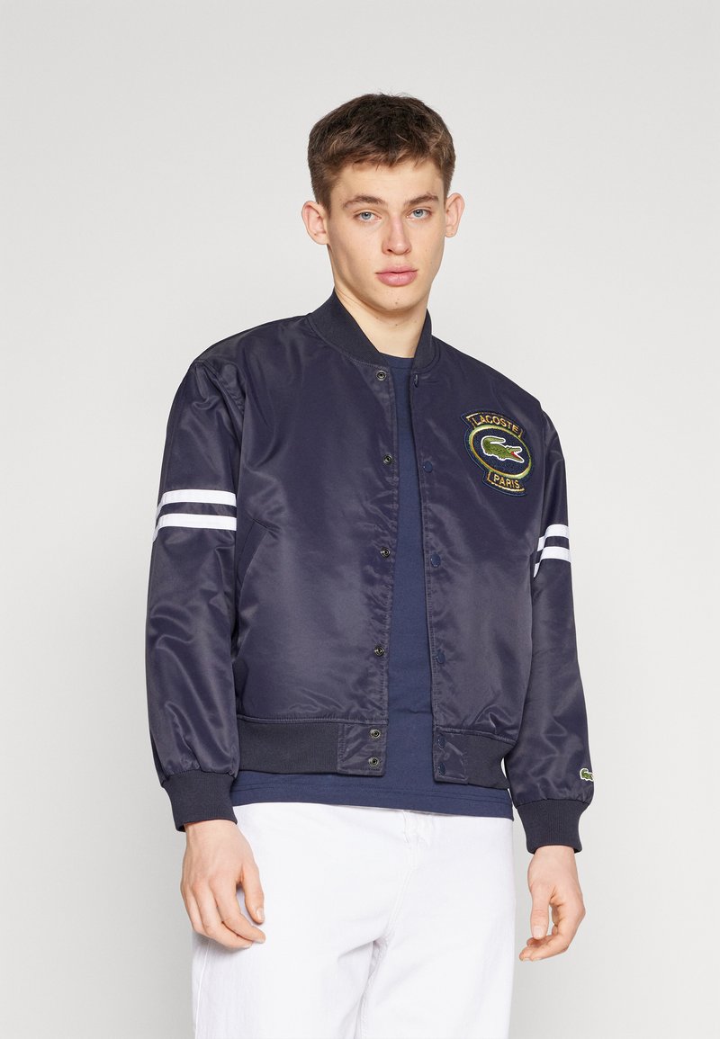 Veste bomber en satin marine avec des manches à rayures blanches, des poignets côtes et un patch logo Lacoste sur la poitrine. Portée par-dessus un haut sombre.